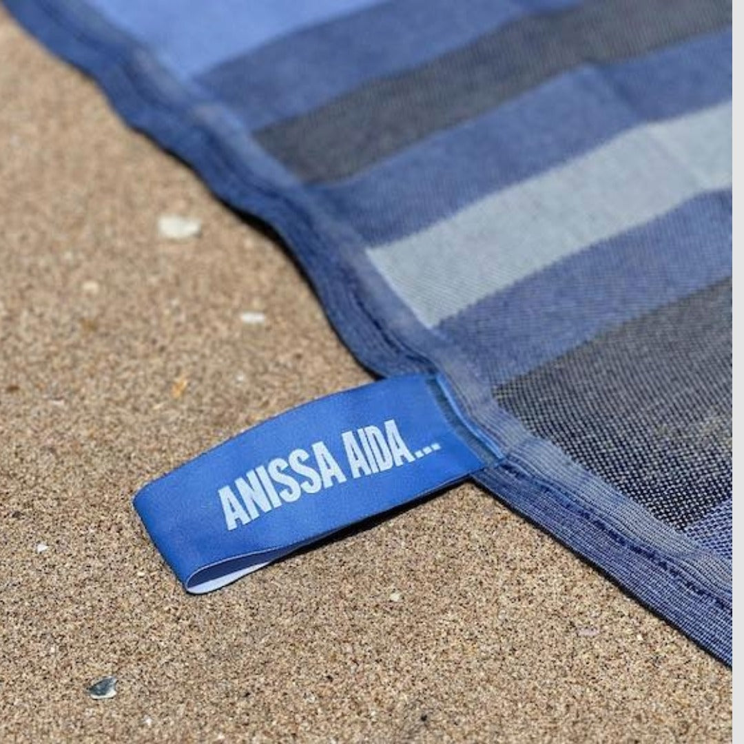 Blue striped towel with 'ANISSA AIDA' label on sand
