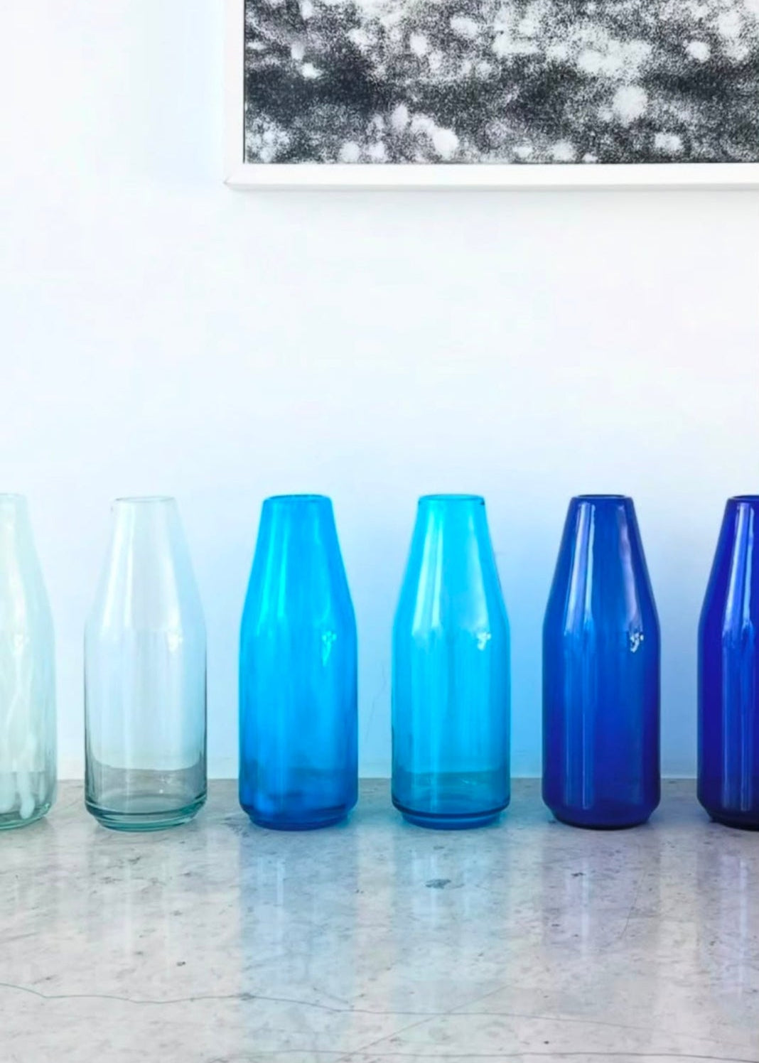 A row of seven translucent glass vases in various shades of blue and clear glass, lined up on a light marble surface against a white wall with a framed abstract print.