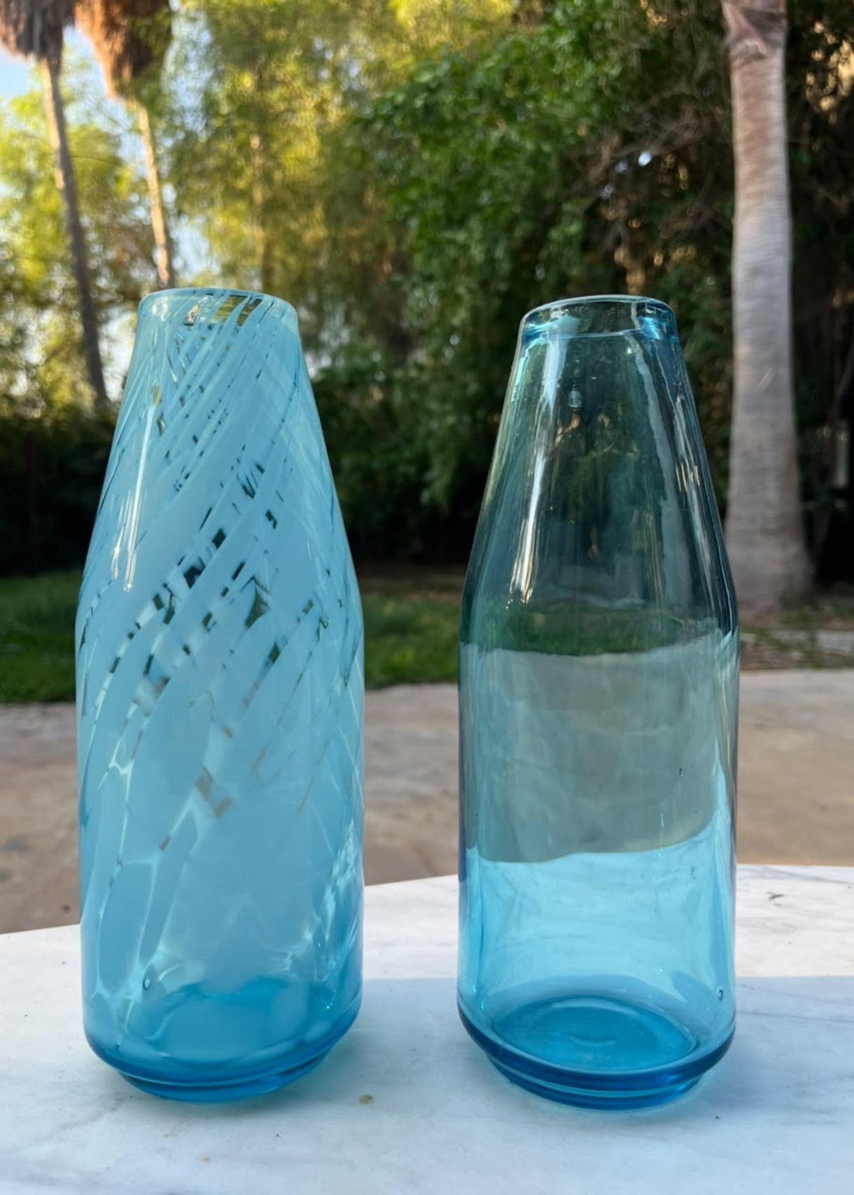 Two light blue glass vases with different textured patterns, standing on a white marble table outdoors with a garden background.