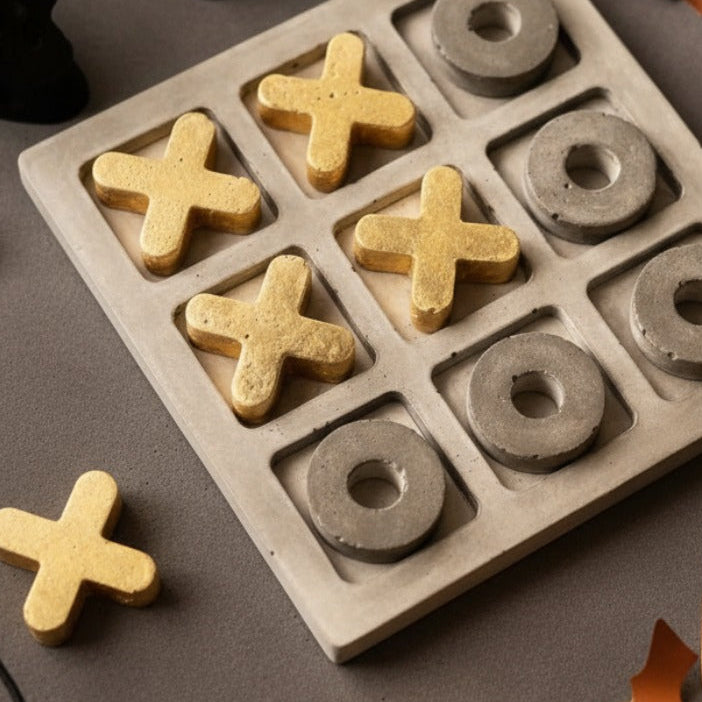 Concrete tic-tac-toe board with gold 'X' and gray 'O' pieces on a dark surface with decorative elements.